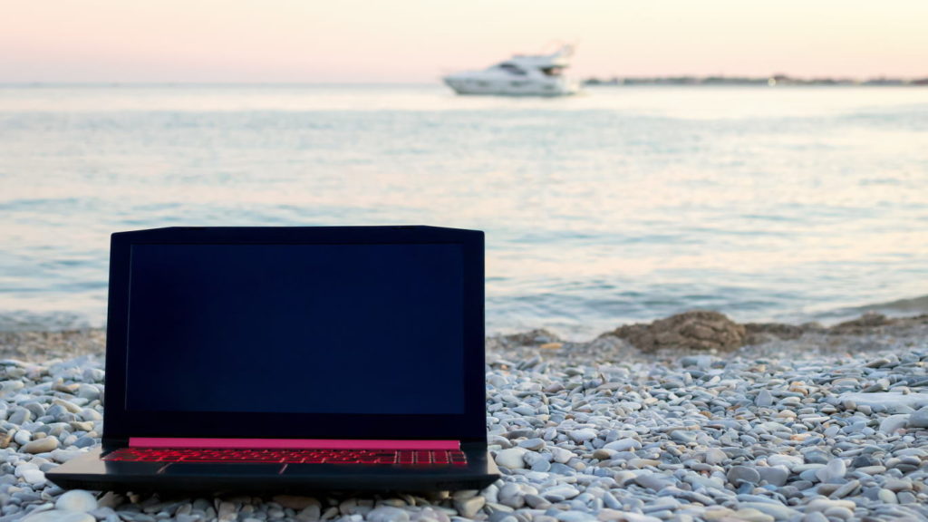Close-up and soft focus of laptop that lies on rocks on the beach by the sea and a blurry white yacht in the background. The concept of working as a freelancer, business and financial independence (Close-up and soft focus of laptop that lies on rocks